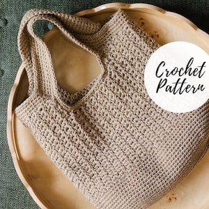 May include: A beige crocheted tote bag with a wide strap, resting on a wooden tray. The bag has a textured appearance and a large opening. A white circle with the words "Crochet Pattern" is on the right side of the bag.