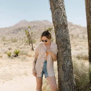 May include: A woman wearing a white crochet cardigan, denim shorts, and white cowboy boots stands next to a tree in a desert landscape.