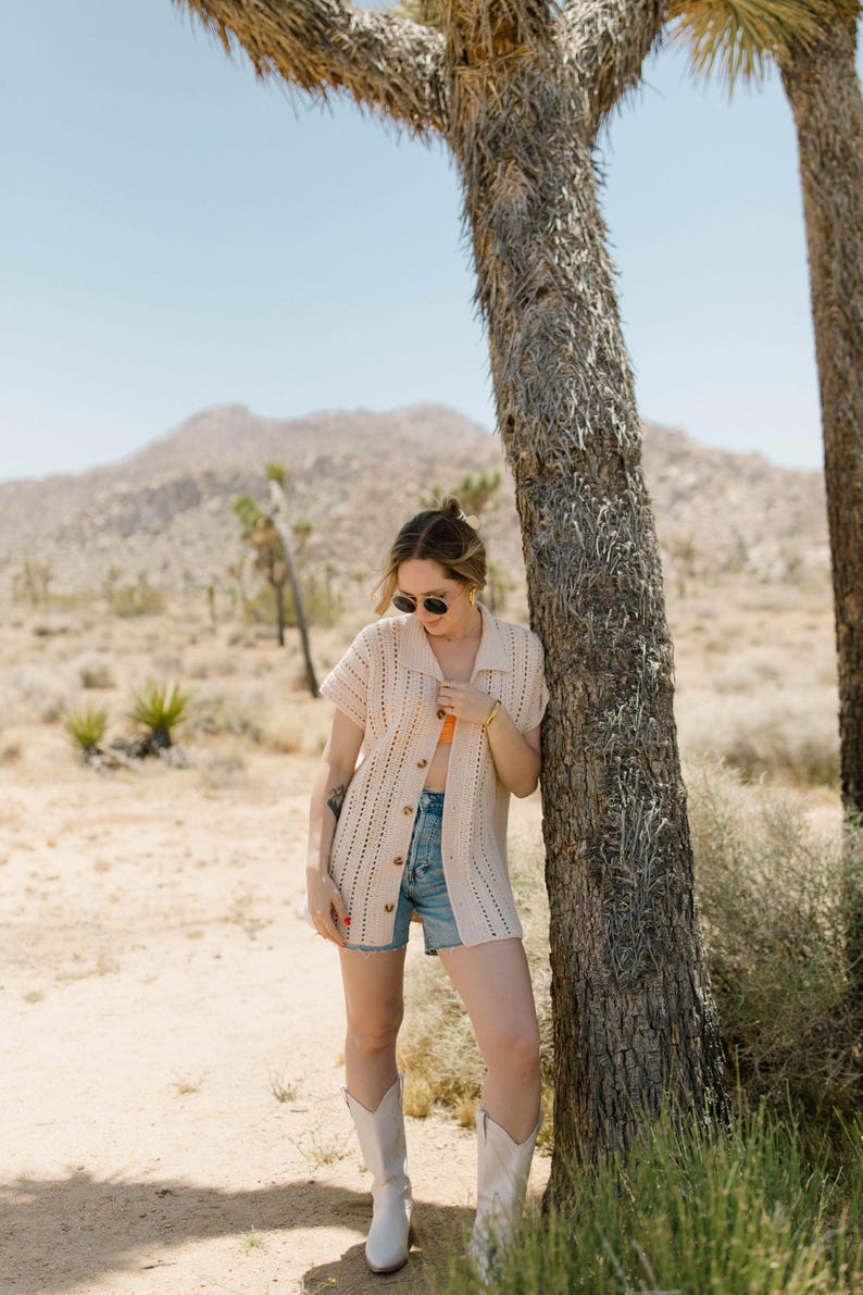 May include: A woman wearing a white crochet cardigan, denim shorts, and white cowboy boots stands next to a tree in a desert landscape.