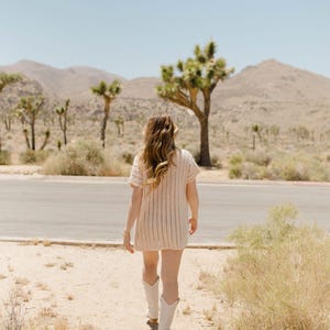 May include: A woman wearing a white crochet dress and white cowboy boots walks on a dirt path in a desert landscape.