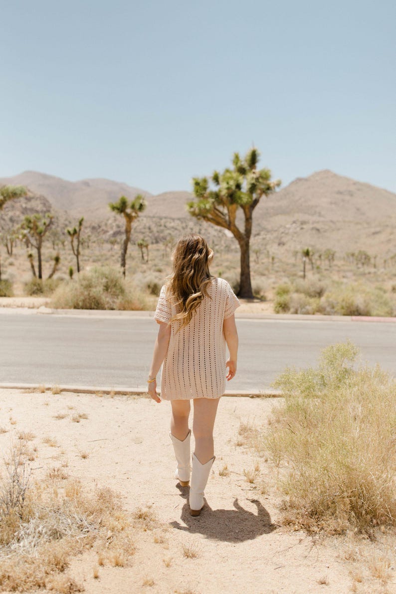 May include: A woman wearing a white crochet dress and white cowboy boots walks on a dirt path in a desert landscape.