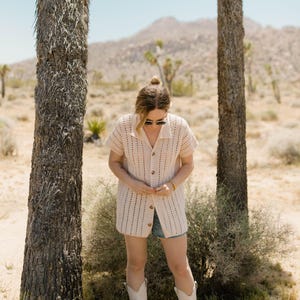 May include: A woman wearing a white crochet cardigan with a button-up front, denim shorts, and white cowboy boots. She is standing in a desert landscape with trees in the background.