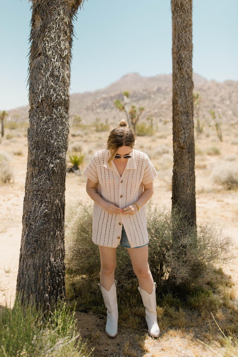 May include: A woman wearing a white crochet cardigan with a button-up front, denim shorts, and white cowboy boots. She is standing in a desert landscape with trees in the background.