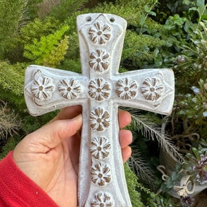May include: A white ceramic cross with a raised floral design. The cross has a distressed white finish with brown accents.