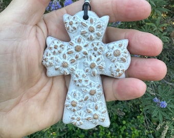 May include: White ceramic cross pendant with a floral design. The cross is decorated with small, raised flowers in a light brown color. The pendant is hanging from a black cord.