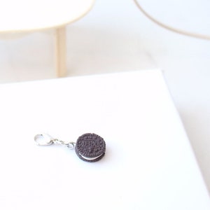 May include: A miniature Oreo cookie charm with a silver-colored clasp. The charm features a detailed replica of the classic chocolate sandwich cookie with white cream filling. The charm is a playful accessory, perfect for jewelry or craft projects.
