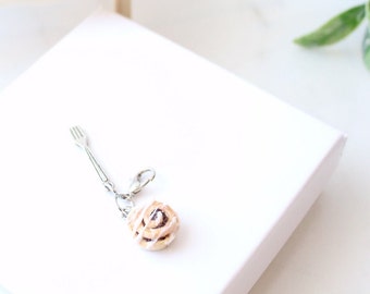 May include: A silver fork charm attached to a silver chain with a small, white-frosted cinnamon roll charm.