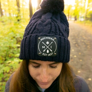 May include: A navy blue knit beanie with a pom-pom on top. The hat features a black square patch with the word "MICHIGAN" and the text "EST 1837" in white. The patch also has a compass design with arrows and a tree.