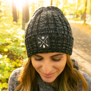 May include: A black and grey knit beanie with a black patch featuring a white snowflake, crossed arrows, and the letters "MI".