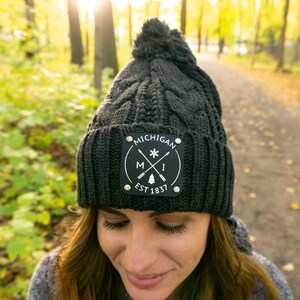 May include: A dark gray cable-knit beanie with a pom-pom on top. The hat features a black patch with the word "MICHIGAN" and the state's initials, along with "EST 1837". The hat is worn outdoors.