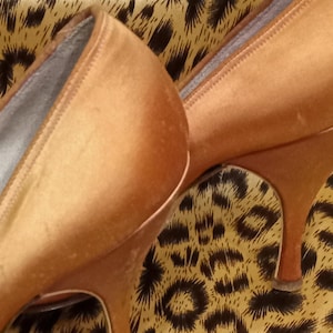 May include: A pair of high-heeled shoes made of a soft, satiny material. The shoes are a light brown color and have a pointed toe. The shoes are sitting on a leopard print fabric.
