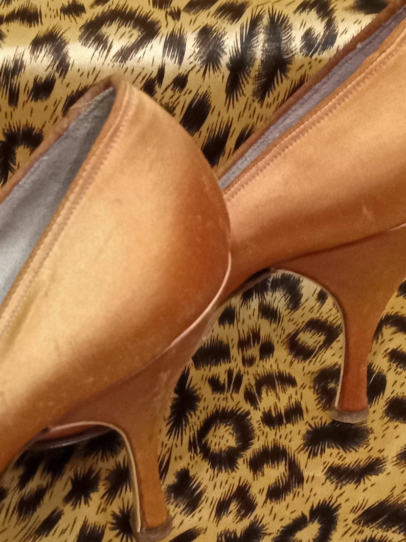May include: A pair of high-heeled shoes made of a soft, satiny material. The shoes are a light brown color and have a pointed toe. The shoes are sitting on a leopard print fabric.