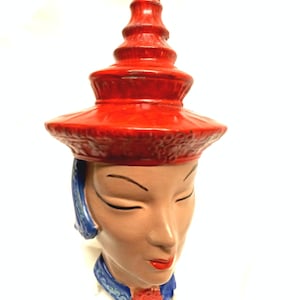 May include: A ceramic head sculpture with closed eyes, red lips, and a red hat. The face is tan with blue accents around the neck and hair. The hat has a tiered design and is also red.