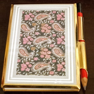 May include: A small, gold-trimmed notebook with a paisley pattern on the cover. A gold pencil with a red eraser is attached to the side.