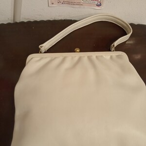 Puede incluir: Un bolso de mano blanco de imitación de cuero con un cierre dorado y una correa blanca.