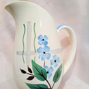 May include: A white ceramic pitcher with a blue and green floral design. The pitcher has a wide mouth and a handle.