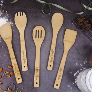 May include: A set of six bamboo kitchen utensils, including a spatula, slotted spoon, and serving spoon, arranged on a dark surface. Each utensil is engraved with "Mom's Kitchen". Flour and nuts are scattered around the utensils.