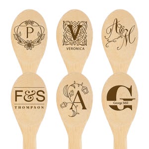 May include: Six wooden spoons with engraved designs. Each spoon features a different design, including initials, names, and decorative elements. The spoons are made of light-colored wood and are ideal for kitchen use.