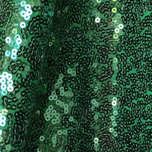 Op de afbeelding: Close-up van een groene paillettenstof. De pailletten zijn een levendig smaragdgroen, vangen het licht op en creëren een glinsterend effect. De stof lijkt gedrapeerd te zijn, met plooien en schaduwen die diepte toevoegen. Ideaal voor kleding of decoratieve projecten.