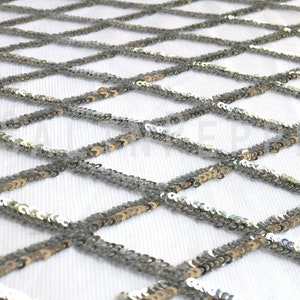 May include: Close-up of a silver sequin fabric with a diamond pattern. The sequins are sewn onto a white mesh backing. The fabric has a reflective quality, catching the light and creating a shimmering effect. Ideal for apparel, decor, and craft projects.