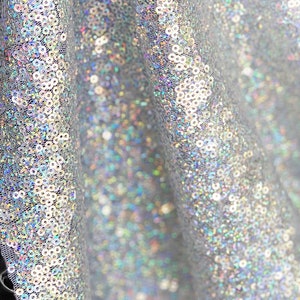 May include: Close-up of a shimmering silver fabric covered in small, round sequins. The fabric is draped, creating soft folds and shadows. The sequins reflect light, creating a sparkling effect. The fabric is likely used for apparel or decorative purposes.