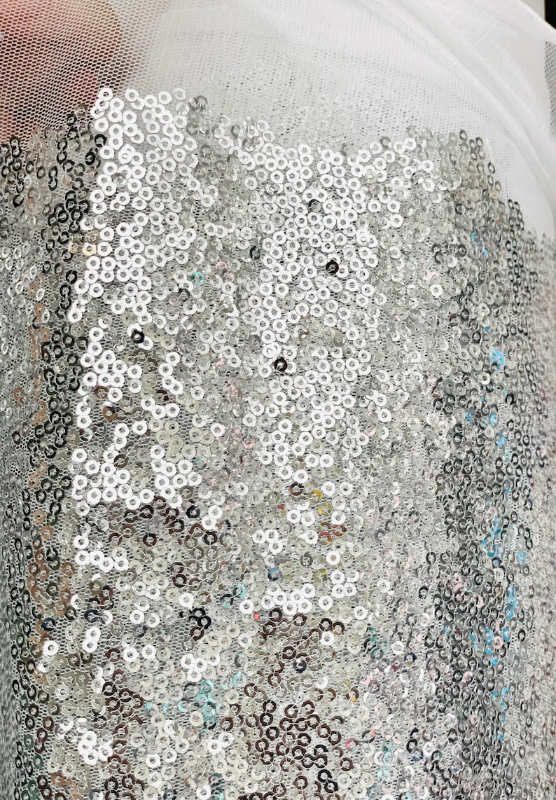 Puede incluir: Primer plano de una tela cubierta de peque&ntilde;as lentejuelas redondas plateadas. Las lentejuelas est&aacute;n densamente empaquetadas, creando un efecto brillante. Una tela blanca transparente es visible en la parte superior, agregando una capa de textura. El efecto general es glamuroso y llamativo.