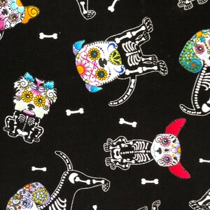 May include: Black fabric featuring a repeating pattern of dogs with sugar skull faces and white skeleton bodies. The dogs have colorful floral designs on their faces and are interspersed with white bone shapes.