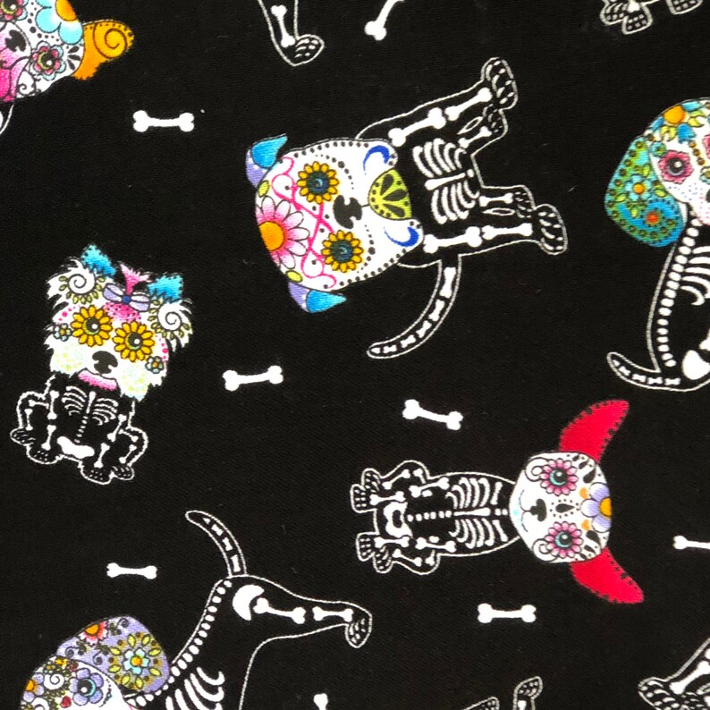 May include: Black fabric featuring a repeating pattern of dogs with sugar skull faces and white skeleton bodies. The dogs have colorful floral designs on their faces and are interspersed with white bone shapes.