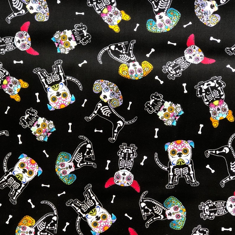 May include: Black fabric with a repeating pattern of dog skeletons decorated with colorful floral designs. White bone shapes are scattered across the background. The dogs are in various poses.