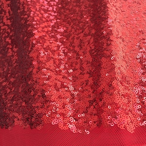 May include: Close-up of a red sequin fabric. The sequins are densely packed and reflect light, creating a shimmering effect. The fabric has a mesh backing, visible at the bottom of the image. The overall texture is smooth and luxurious.