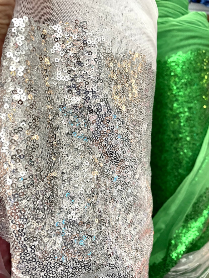 Puede incluir: Primer plano de un rollo de tela de lentejuelas plateadas junto a un rollo de tela de lentejuelas verdes. La tela plateada est&aacute; densamente cubierta de peque&ntilde;as lentejuelas redondas, que reflejan la luz. La tela verde tambi&eacute;n est&aacute; cubierta de lentejuelas.