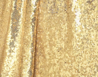 May include: Close-up of a gold sequin fabric. The fabric is densely covered in small, round, shiny sequins. The sequins reflect light, creating a shimmering effect. The fabric is draped, with folds and shadows adding depth.
