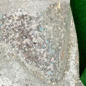 Puede incluir: Primer plano de una tela con lentejuelas plateadas. Las lentejuelas est&aacute;n densamente empaquetadas y reflejan la luz, creando un efecto brillante. La tela es de color claro, posiblemente blanca o plateada, y probablemente se utiliza para ropa o decoraci&oacute;n.