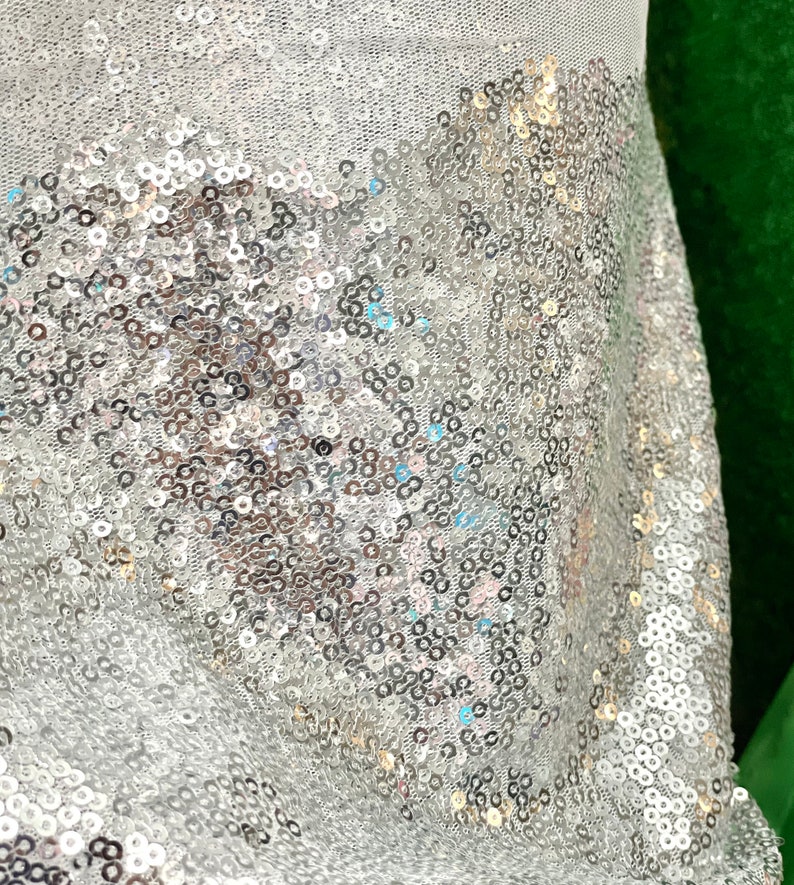 Puede incluir: Primer plano de una tela con lentejuelas plateadas. Las lentejuelas est&aacute;n densamente empaquetadas y reflejan la luz, creando un efecto brillante. La tela es de color claro, posiblemente blanca o plateada, y probablemente se utiliza para ropa o decoraci&oacute;n.
