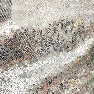 Puede incluir: Primer plano de una tela brillante cubierta de lentejuelas plateadas. Las lentejuelas son redondas y densamente empaquetadas, creando una superficie reflectante. La tela tiene un respaldo de malla, agregando textura y dimensi&oacute;n al dise&ntilde;o. Ideal para ropa y manualidades.
