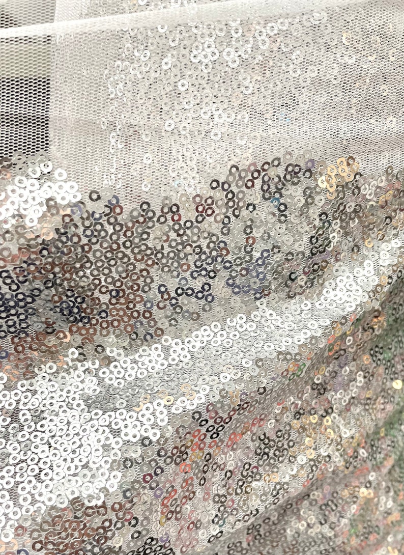 Puede incluir: Primer plano de una tela brillante cubierta de lentejuelas plateadas. Las lentejuelas son redondas y densamente empaquetadas, creando una superficie reflectante. La tela tiene un respaldo de malla, agregando textura y dimensi&oacute;n al dise&ntilde;o. Ideal para ropa y manualidades.