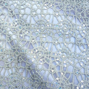 May include: Close-up of a silver lace fabric with a delicate, openwork design. The fabric features a network of interconnected lines and small, shimmering sequins, creating a textured and elegant appearance. The overall effect is a light, airy, and decorative textile.