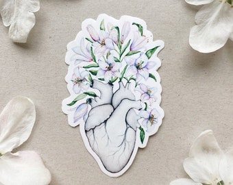 Op de afbeelding: Een wit anatomisch hart met een boeket witte bloemen die eruit groeien. De bloemen zijn geschilderd in aquarel met paarse en groene accenten.