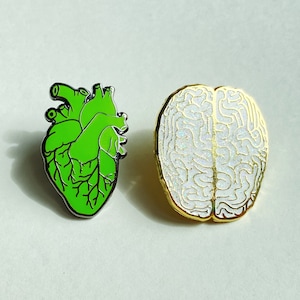 Pode incluir: Dois pinos de esmalte, um com a forma de um coração humano verde e o outro com a forma de um cérebro humano branco.