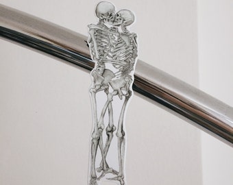 May include: Two skeletons are embracing and kissing. The skeletons are drawn in black and white and are attached to a silver railing.