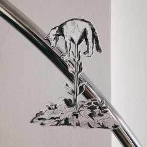 K&ouml;nnte beinhalten: Schwarzwei&szlig;-Illustration eines Hundes, der auf einer Pflanze mit Bl&auml;ttern steht. Die Illustration befindet sich auf einer silbernen Metalloberfl&auml;che.