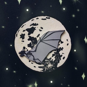 May include: A black and white enamel pin of a bat flying in front of a moon with a textured surface. The bat has a skeletal design and is flying with its wings spread.