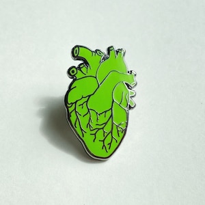 May include: A green anatomical heart-shaped enamel pin with black vein detailing and a silver border. The pin is a stylized representation of a human heart, suitable for medical professionals or those with an interest in anatomy.
