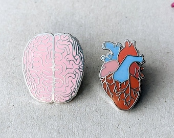 May include: Two enamel pins, one depicting a pink human brain and the other a red, blue, and pink human heart.