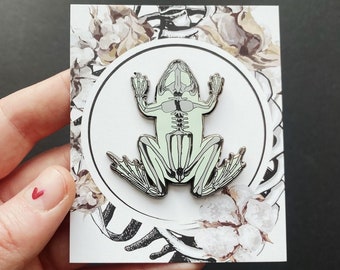 Puede incluir: Un pin de esmalte blanco con una rana esquelética detallada con un cuerpo translúcido verde claro. El pin se muestra en una tarjeta blanca con un diseño floral.
