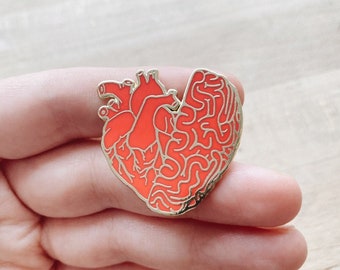 May include: A gold-colored enamel pin shaped like a human heart with a red and white brain design inside.