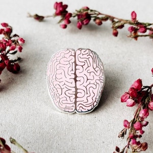 May include: A pink enamel pin shaped like a human brain. The pin is surrounded by pink flowers.