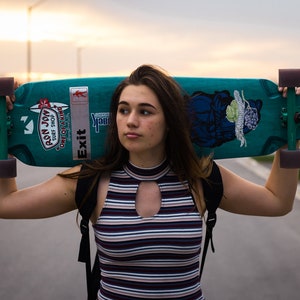 K&ouml;nnte beinhalten: T&uuml;rkisfarbenes Longboard mit lila Rollen und gr&uuml;nen Achsen. Das Board hat Aufkleber und ein grafisches Design. Die Person tr&auml;gt ein gestreiftes Oberteil und einen Rucksack. Das Wort "Exit" ist auf dem Board zu sehen.