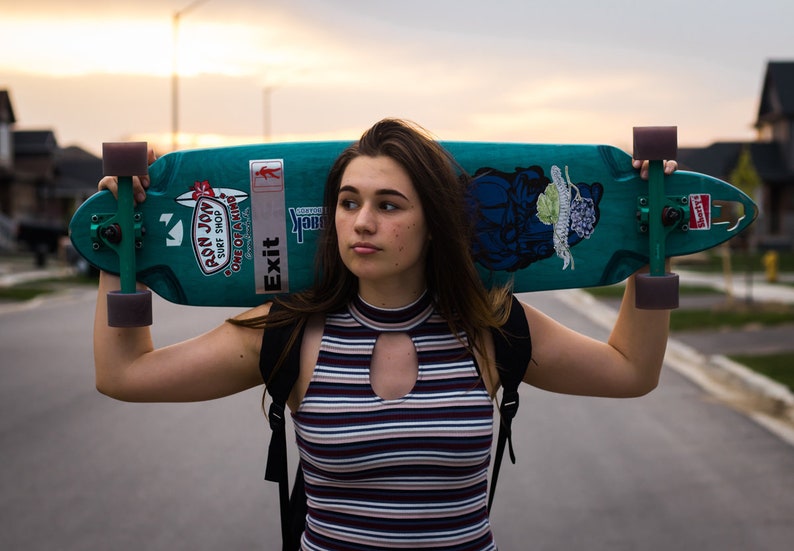 K&ouml;nnte beinhalten: T&uuml;rkisfarbenes Longboard mit lila Rollen und gr&uuml;nen Achsen. Das Board hat Aufkleber und ein grafisches Design. Die Person tr&auml;gt ein gestreiftes Oberteil und einen Rucksack. Das Wort "Exit" ist auf dem Board zu sehen.