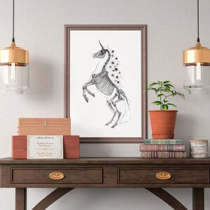 May include: A black and white illustration of a skeletal unicorn with purple flowers in its mane, framed in a brown wood frame and hanging on a white wall above a wooden table with a potted plant, books, and a wooden box.